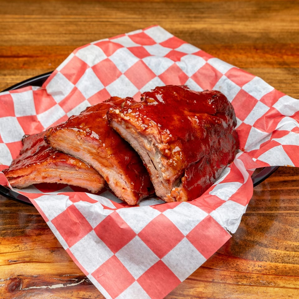 Three saucy BBQ ribs on red and white checkered paper, served on a dark plate.