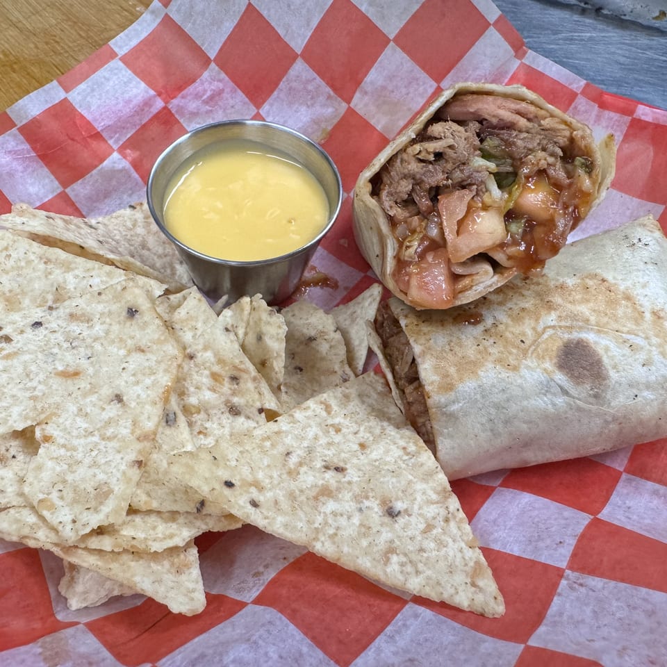 BBQ burrito with pulled pork, a side of tortilla chips, and a cup of cheese dip on checkered paper.