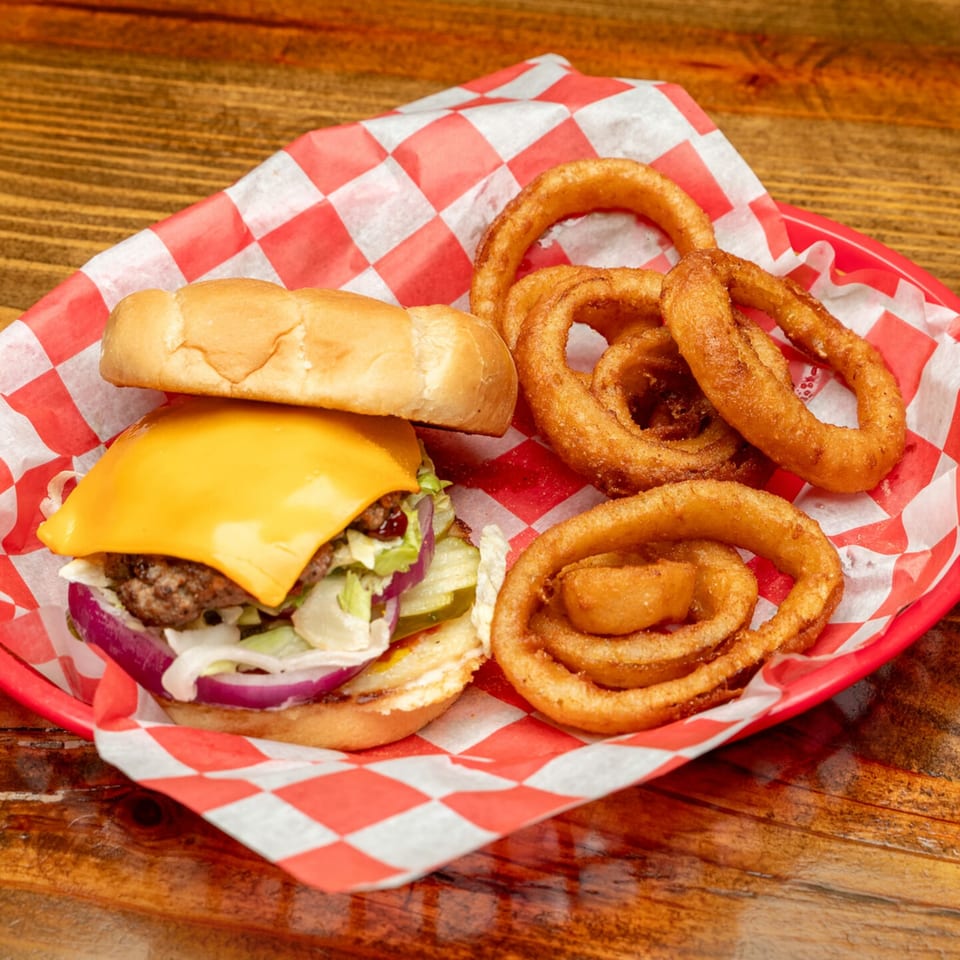 Cheeseburger with lettuce, onion, and pickles, served with crispy onion rings in a red basket.