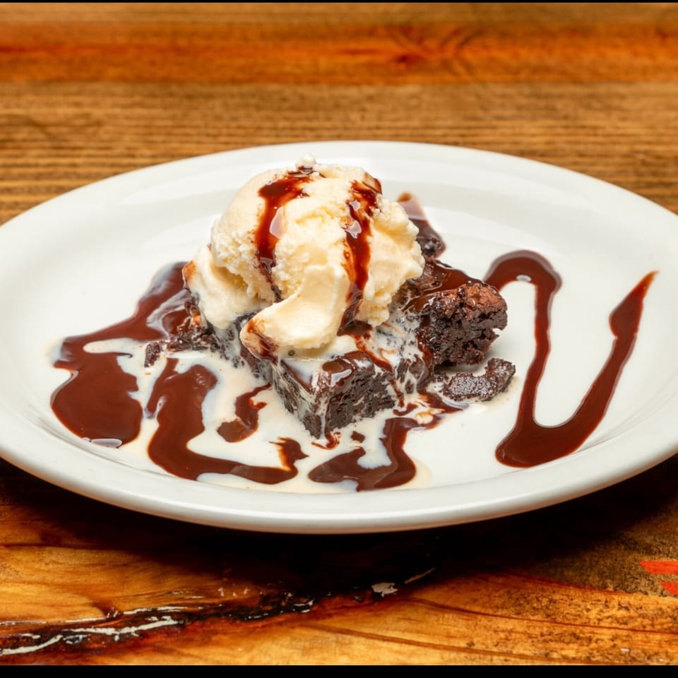 Brownie topped with vanilla ice cream, chocolate syrup, and white sauce on a white plate.